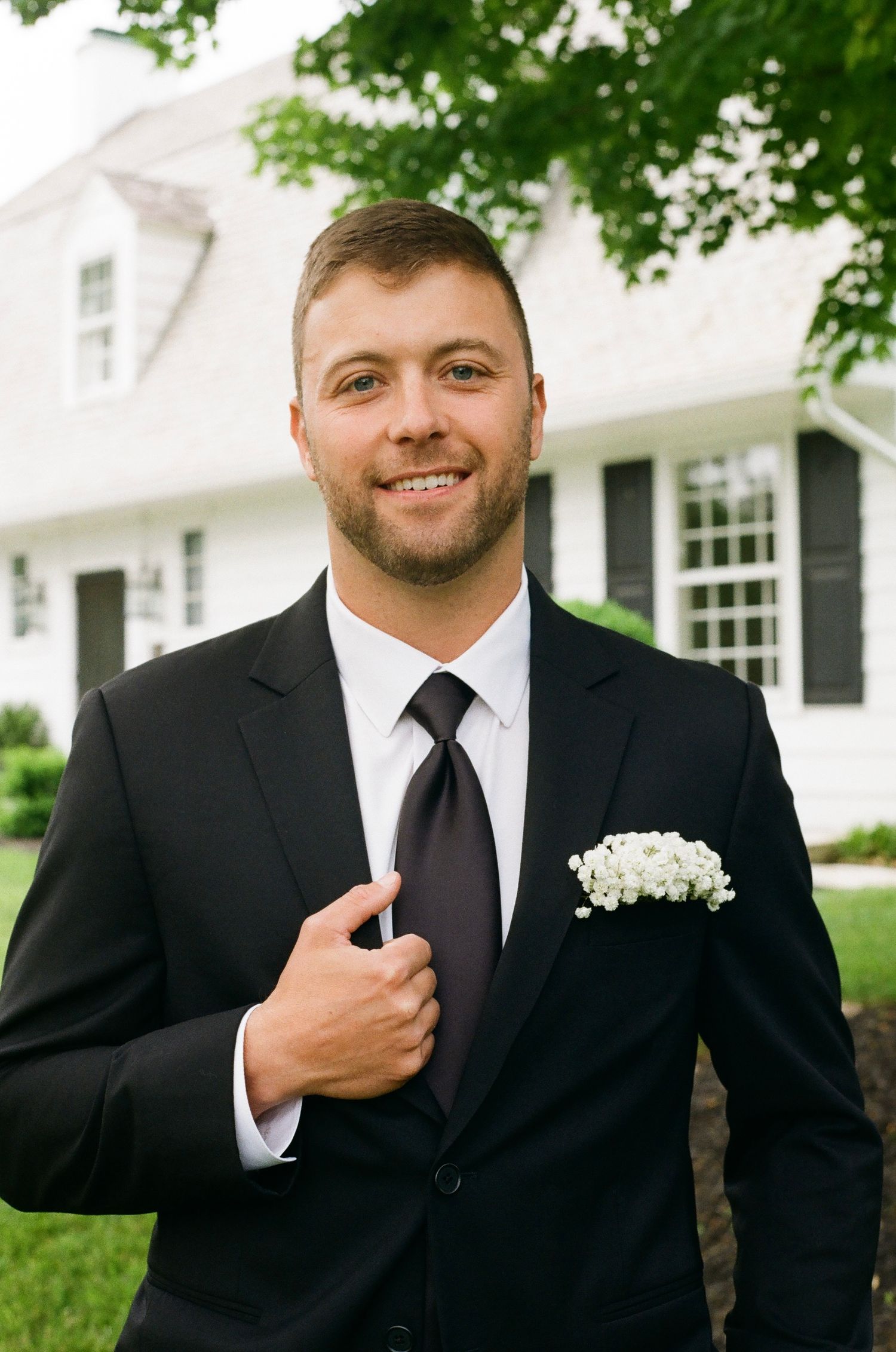 Smiling groom in black suit with white pocket square and dark tie poses outdoors near white house with black shutters.