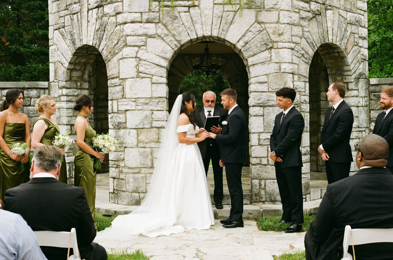 Bride and groom exchange vows at outdoor stone chapel wedding ceremony with bridal party in green and black attire.