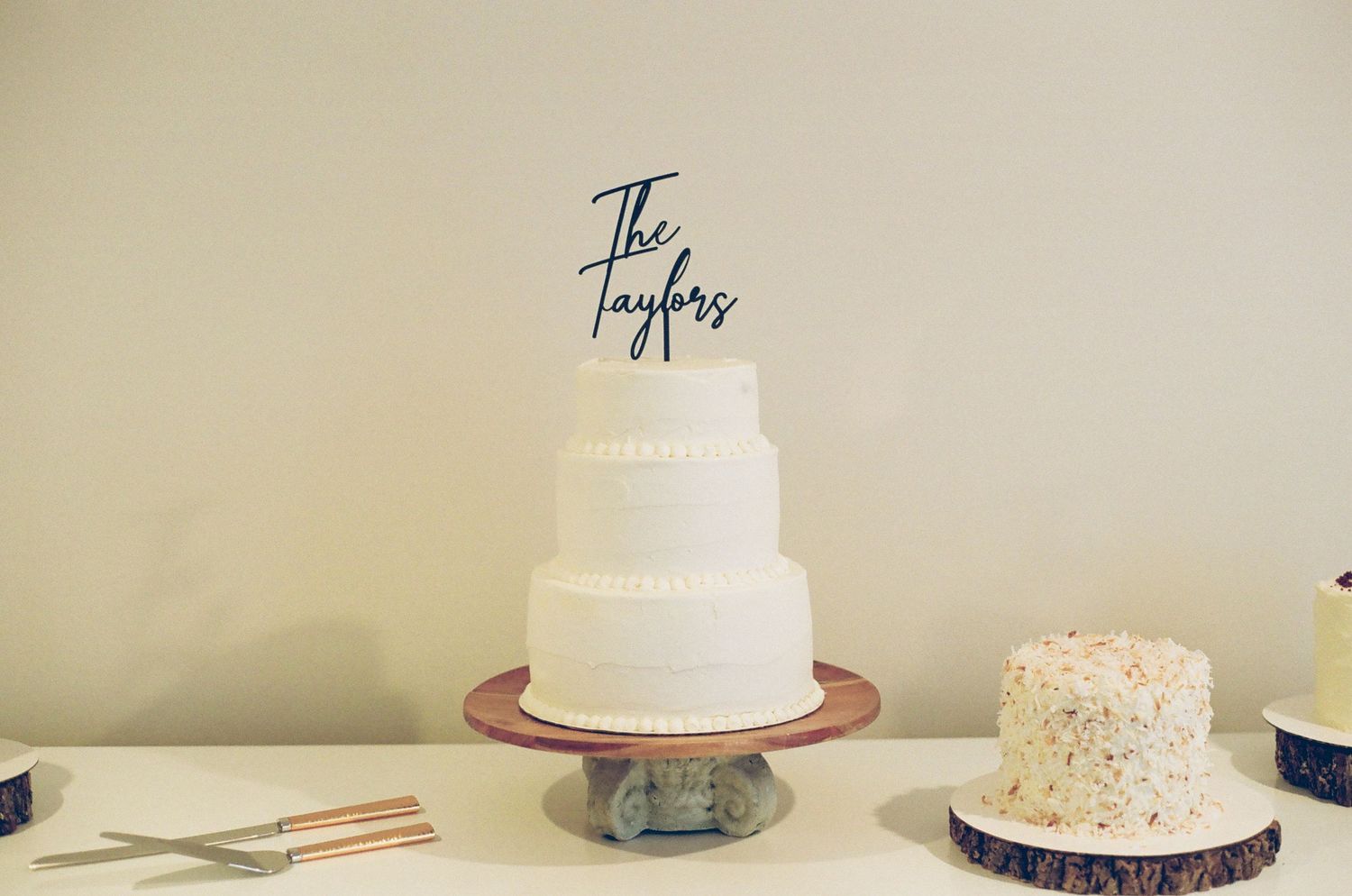 White three-tier wedding cake with The Taylors cake topper on wooden stand at elegant wedding reception.