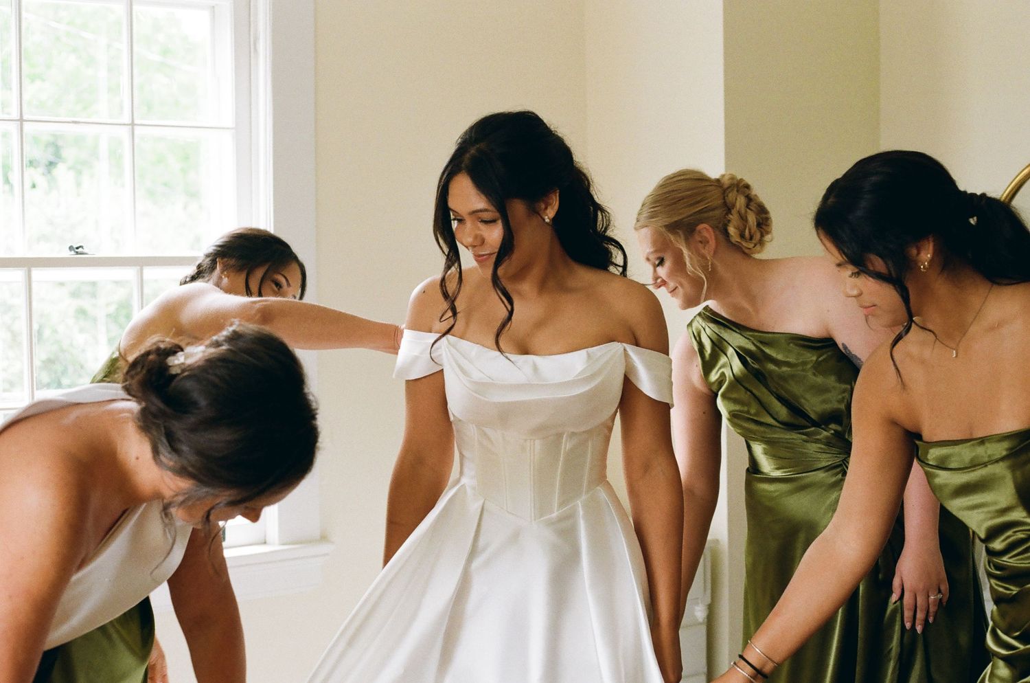Bride in white off-shoulder gown surrounded by bridesmaids in olive green dresses being helped to prepare.