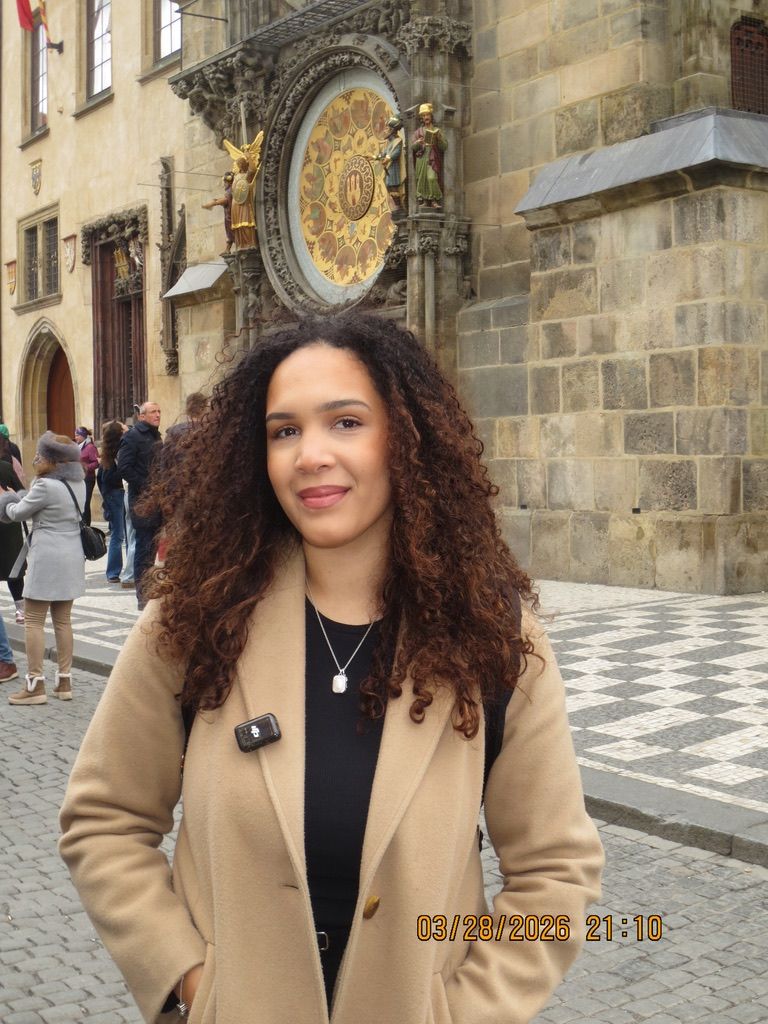 Woman with curly hair in beige coat stands before Prague's Astronomical Clock in Old Town Square.