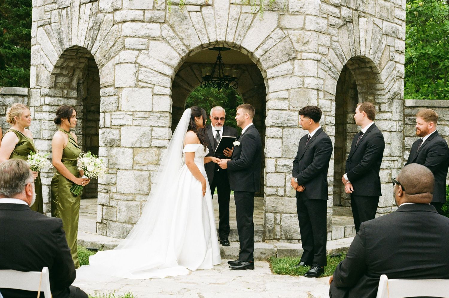Outdoor wedding ceremony at a stone chapel with bride in white gown, groom, officiant, and wedding party.