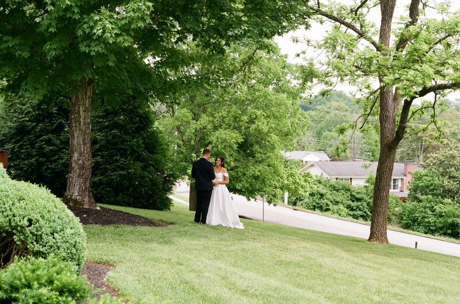 A bride and groom share a romantic moment on a lush green lawn surrounded by tall trees at an outdoor wedding venue.