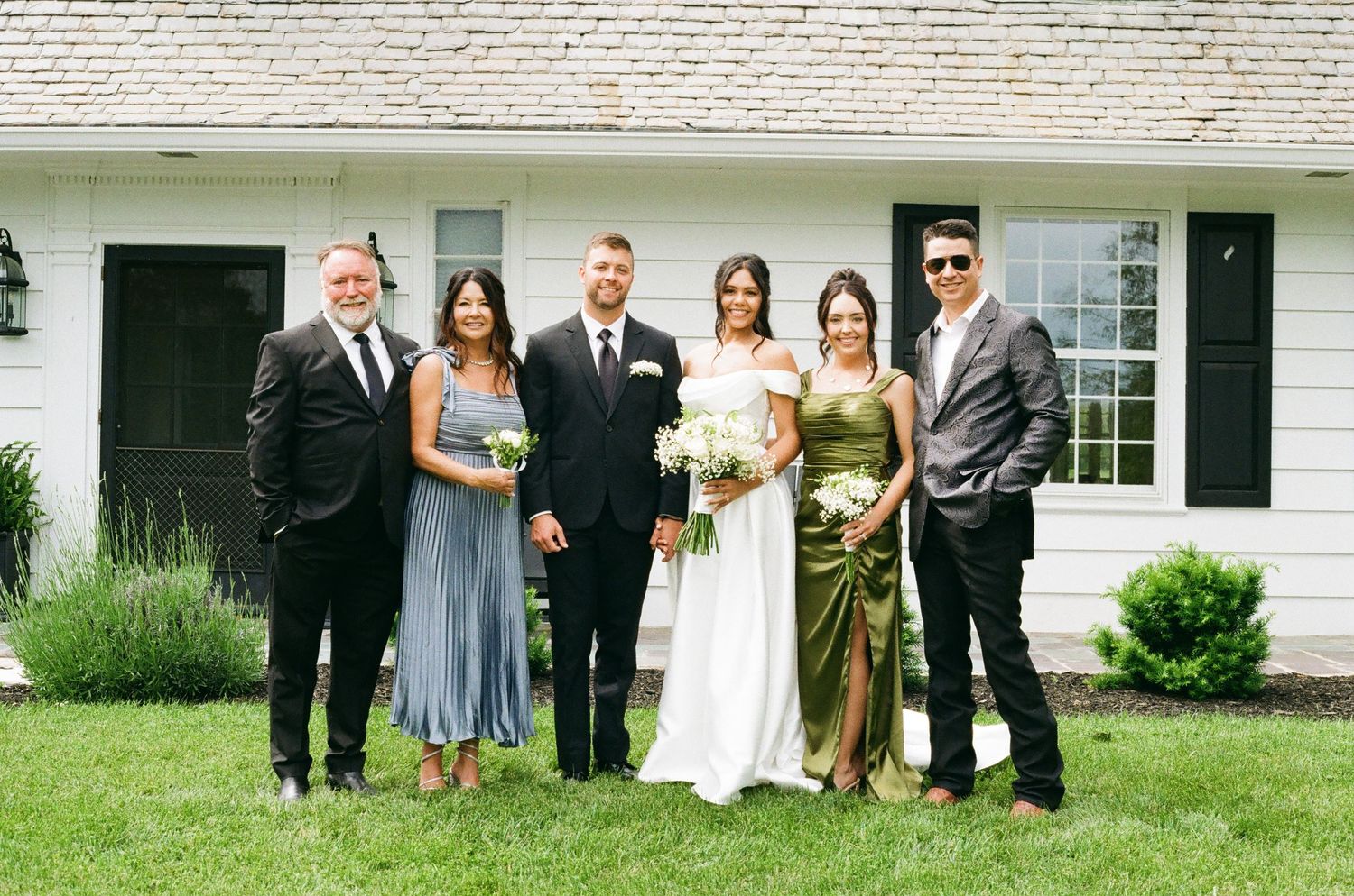 Wedding party posing outdoors in front of white farmhouse, bride in white gown with bouquet, guests in formal attire.