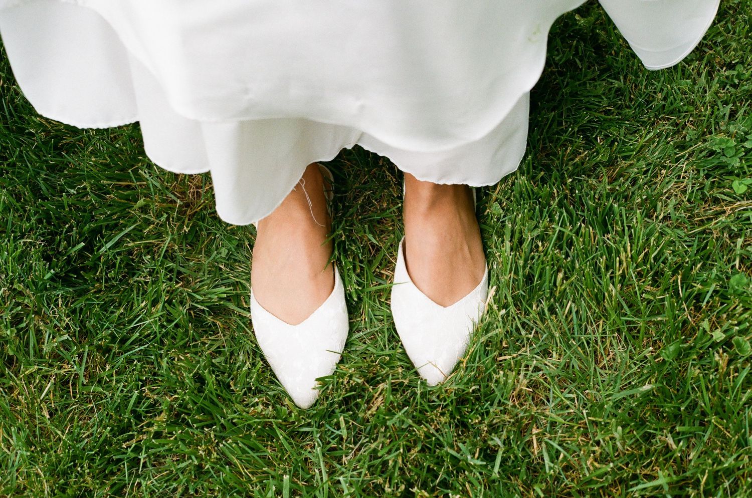 Bride wearing white pointed-toe flats standing on lush green grass beneath a flowing wedding dress.