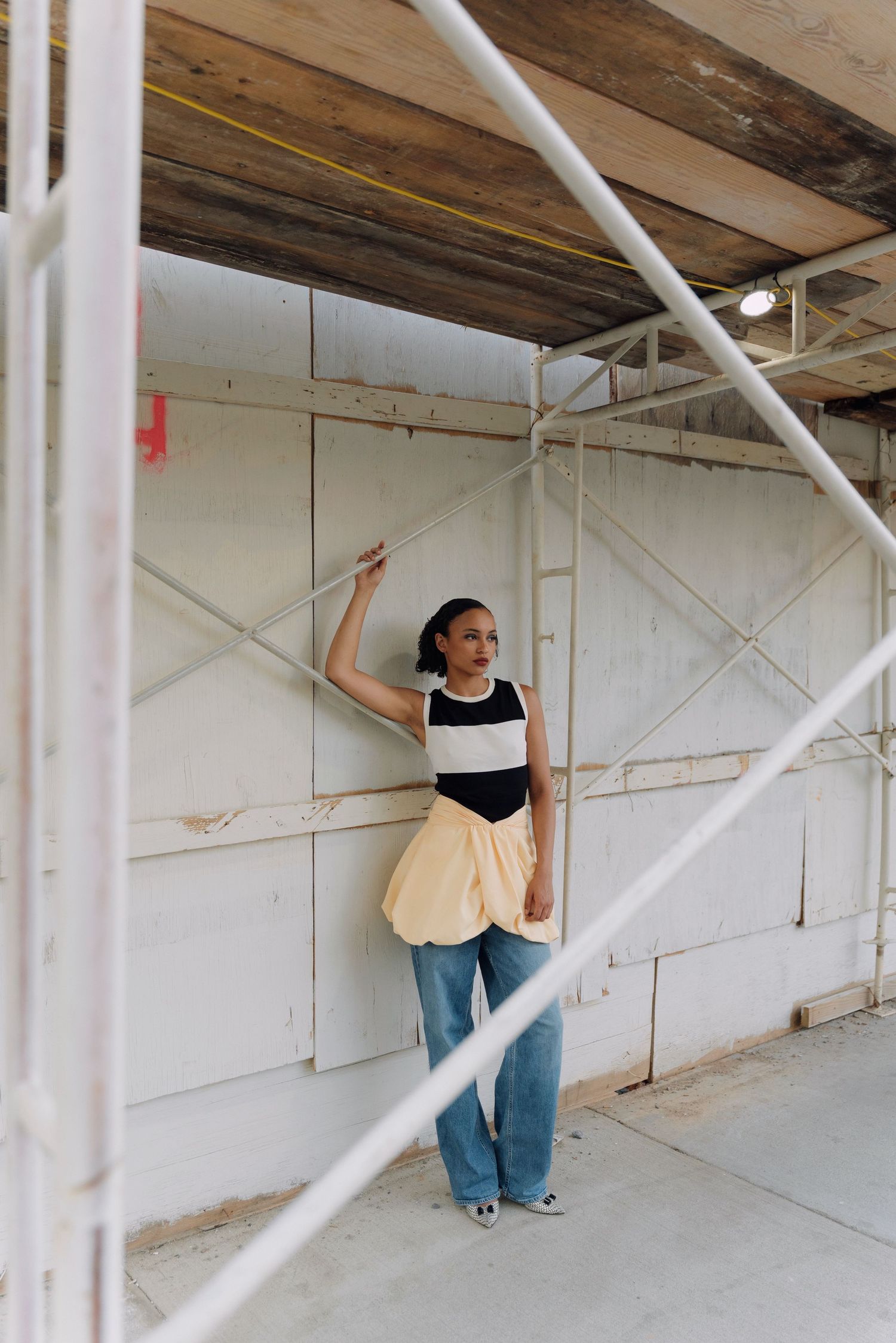 Woman in cream bubble skirt and jeans poses against white wall on construction scaffolding.