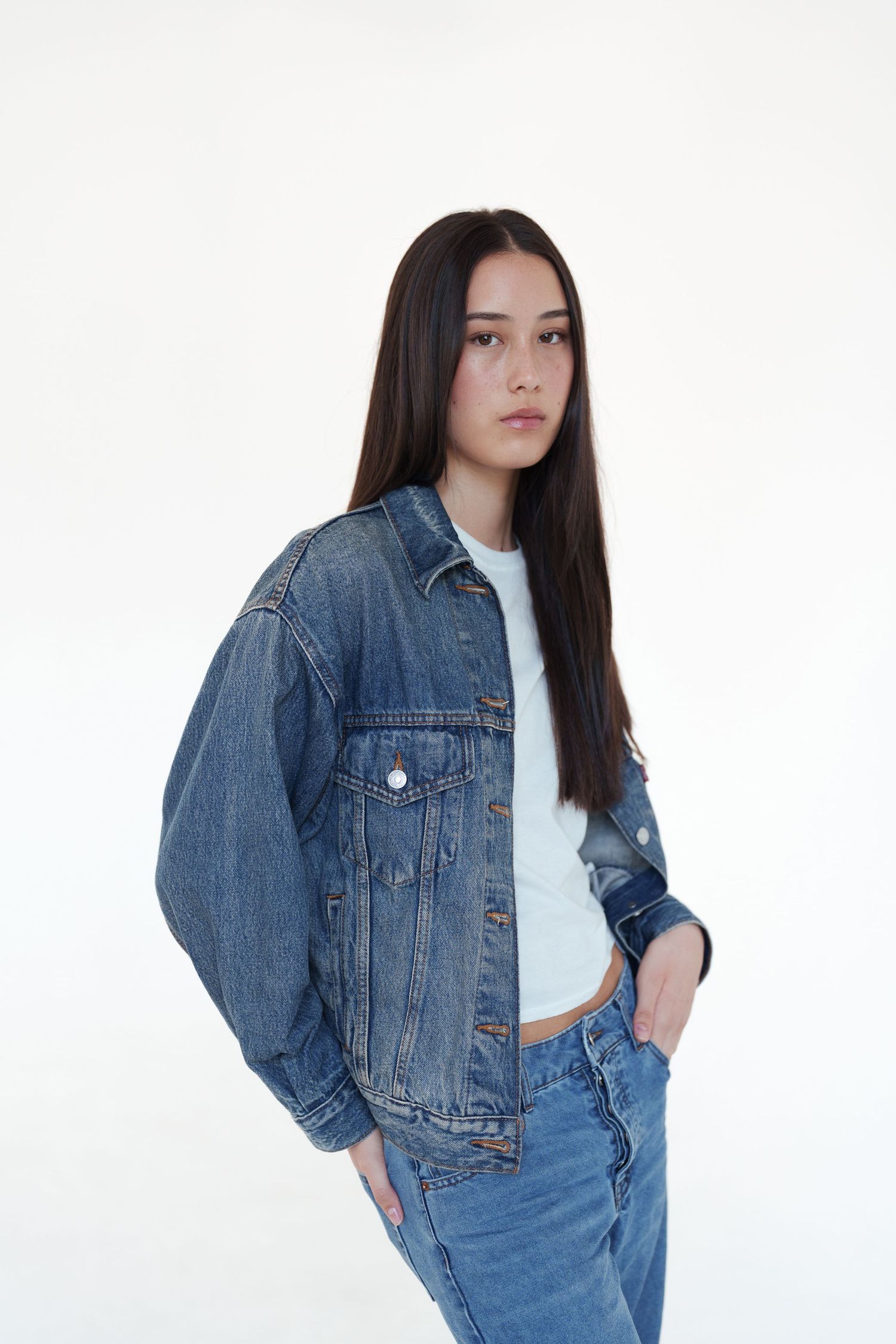 Young woman in blue denim jacket and jeans posing against white background.