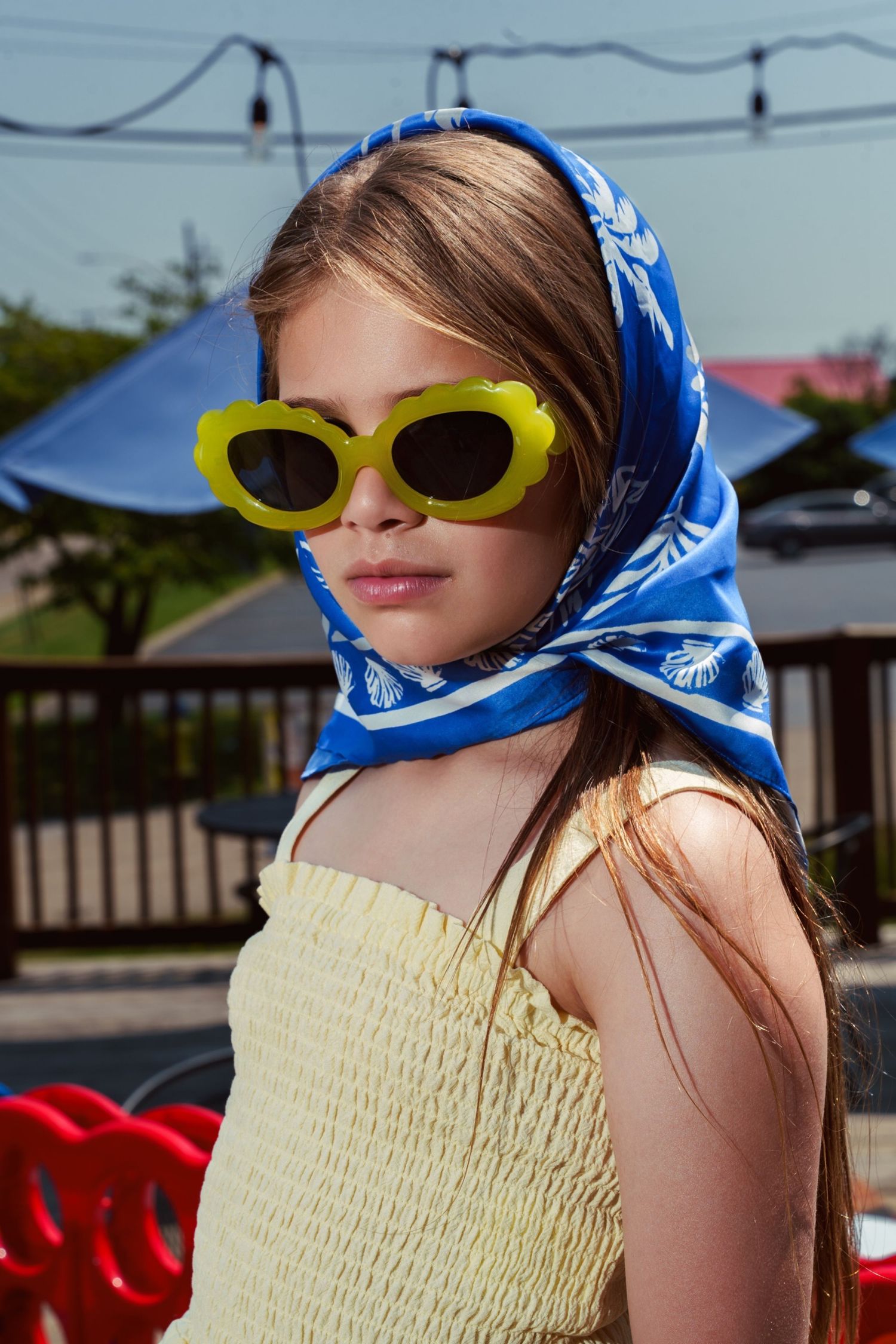 Person wearing yellow oval sunglasses, blue patterned headscarf and yellow smocked top in outdoor setting.