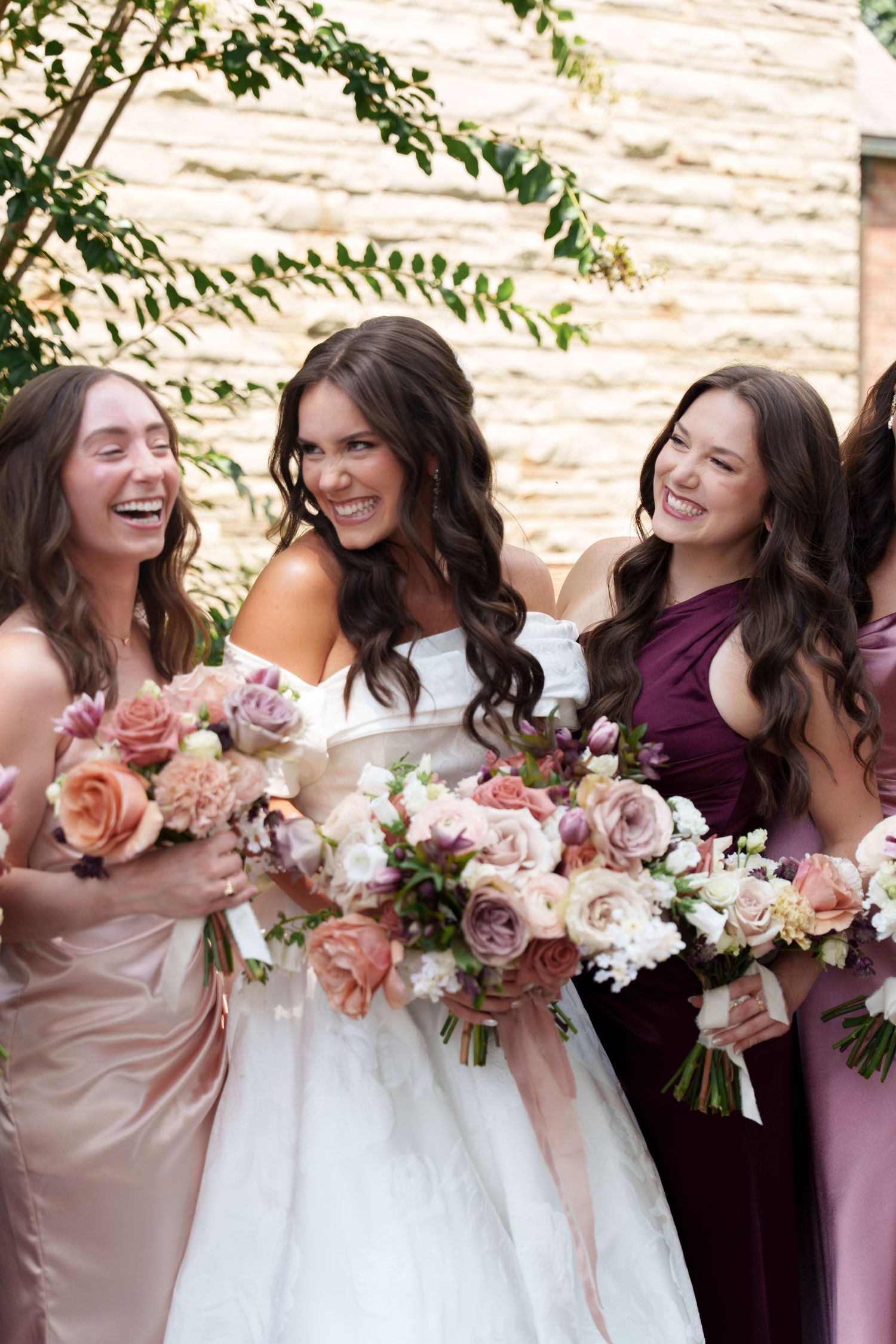 Bridesmaids in burgundy and blush dresses laugh together while holding floral bouquets.