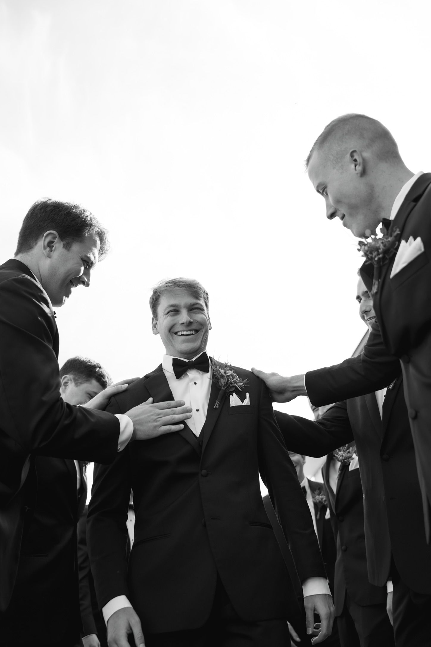 Black and white photo of a smiling groom in a tuxedo being congratulated by groomsmen at a wedding.