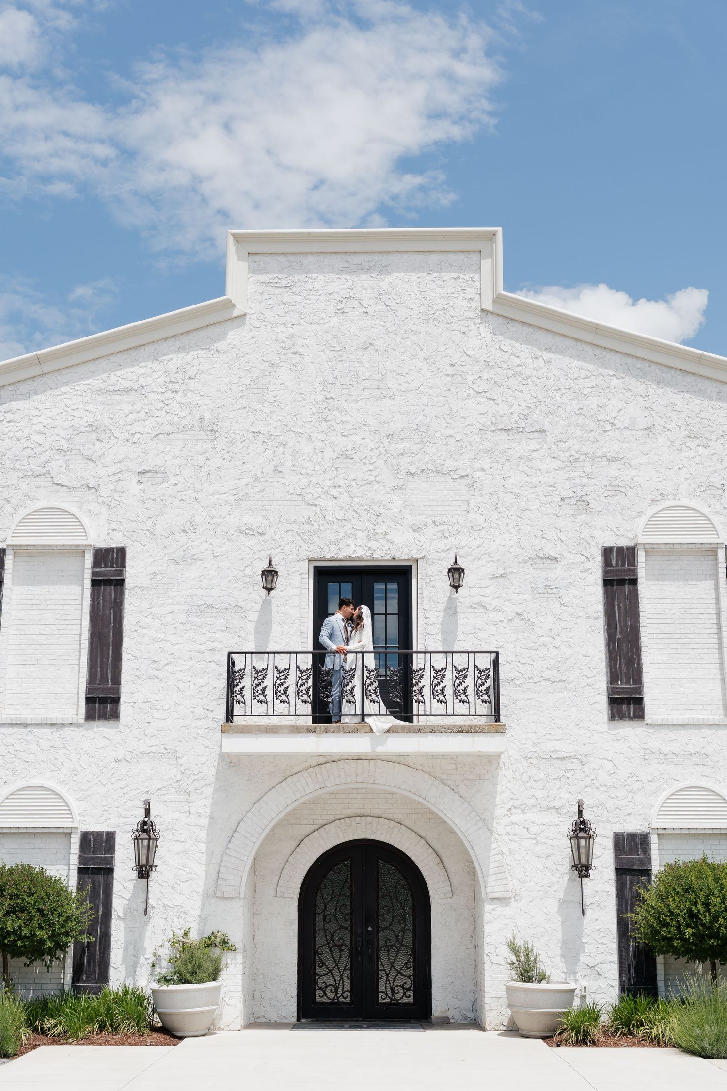 Couple embracing on balcony of elegant white brick wedding venue with arched doorway and dark shutters.