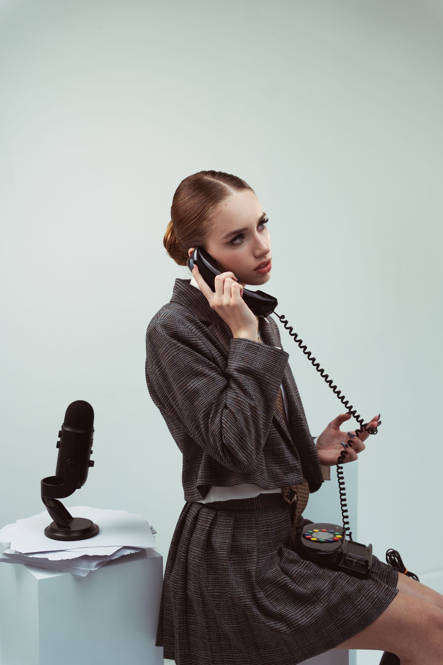 Model wearing dark gray business suit speaking on vintage corded telephone in retro office setting.
