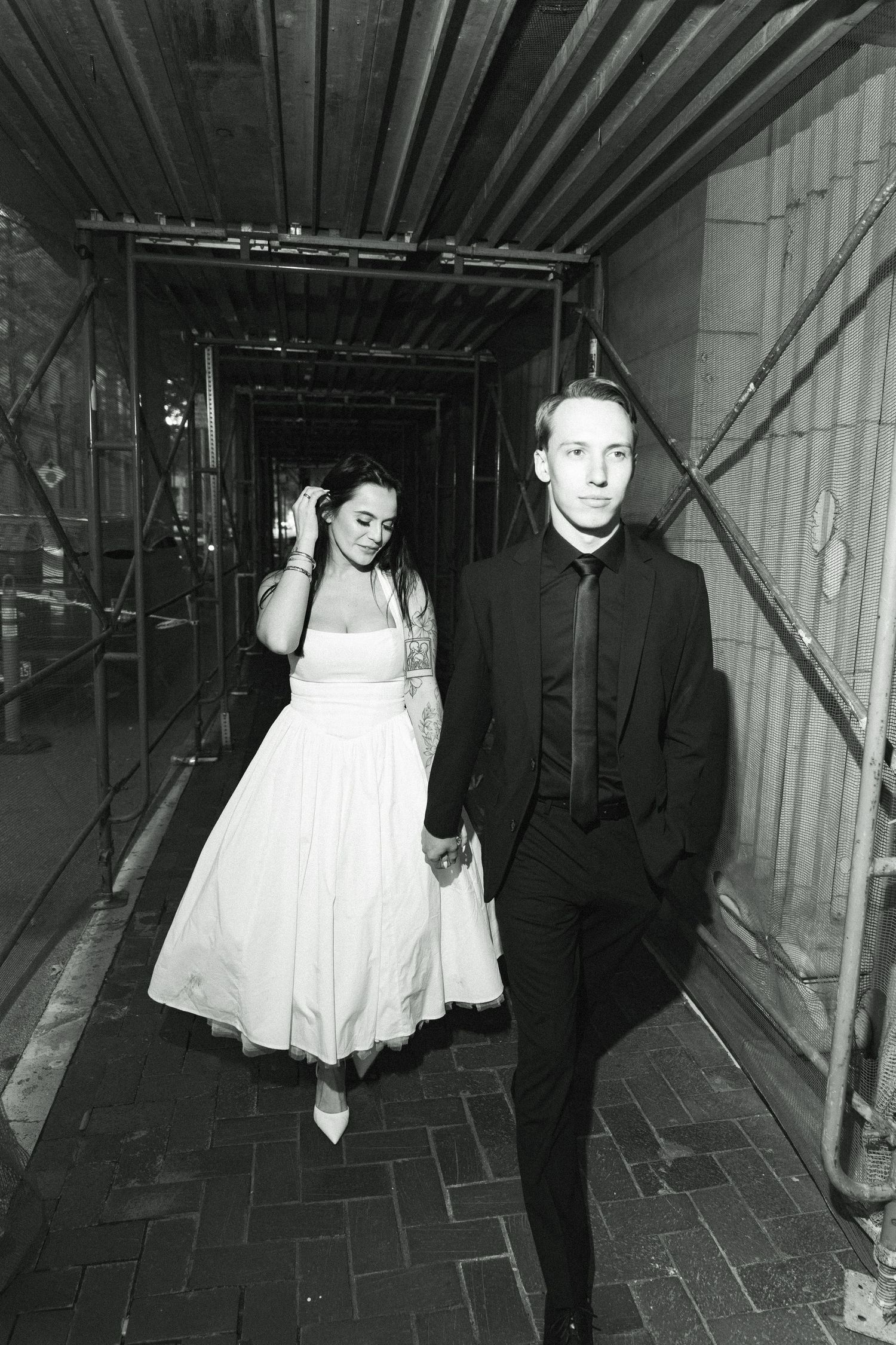 Black and white photo of a bride in white dress and groom in black suit walking hand in hand through a scaffolded corridor.