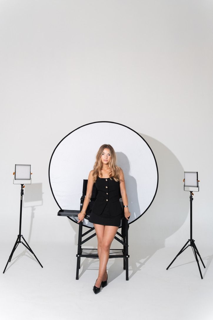 Blonde woman in black mini dress sitting on director chair with studio lighting equipment.