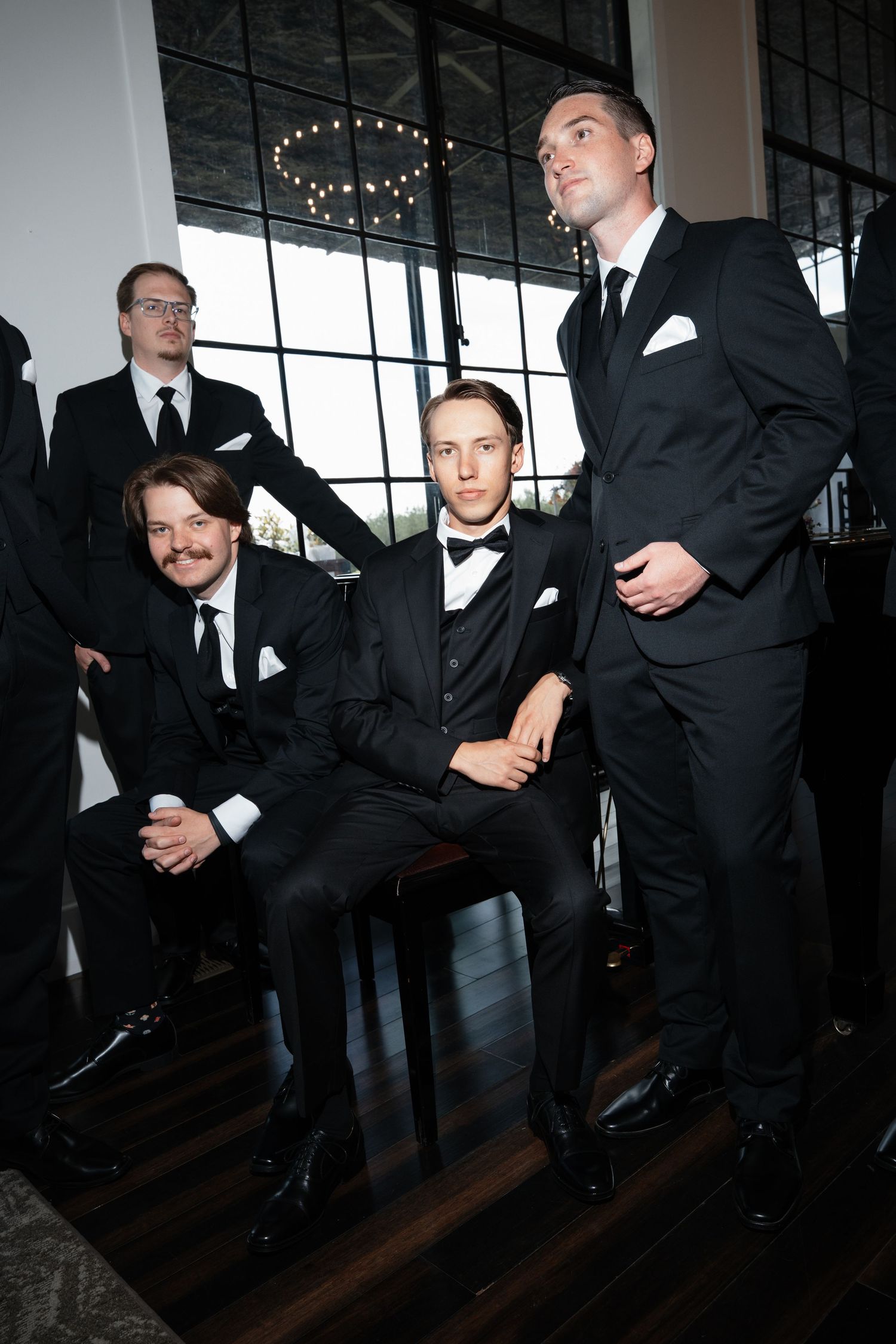 Four men in black suits and bow ties pose formally in an elegant venue with dark hardwood floors.
