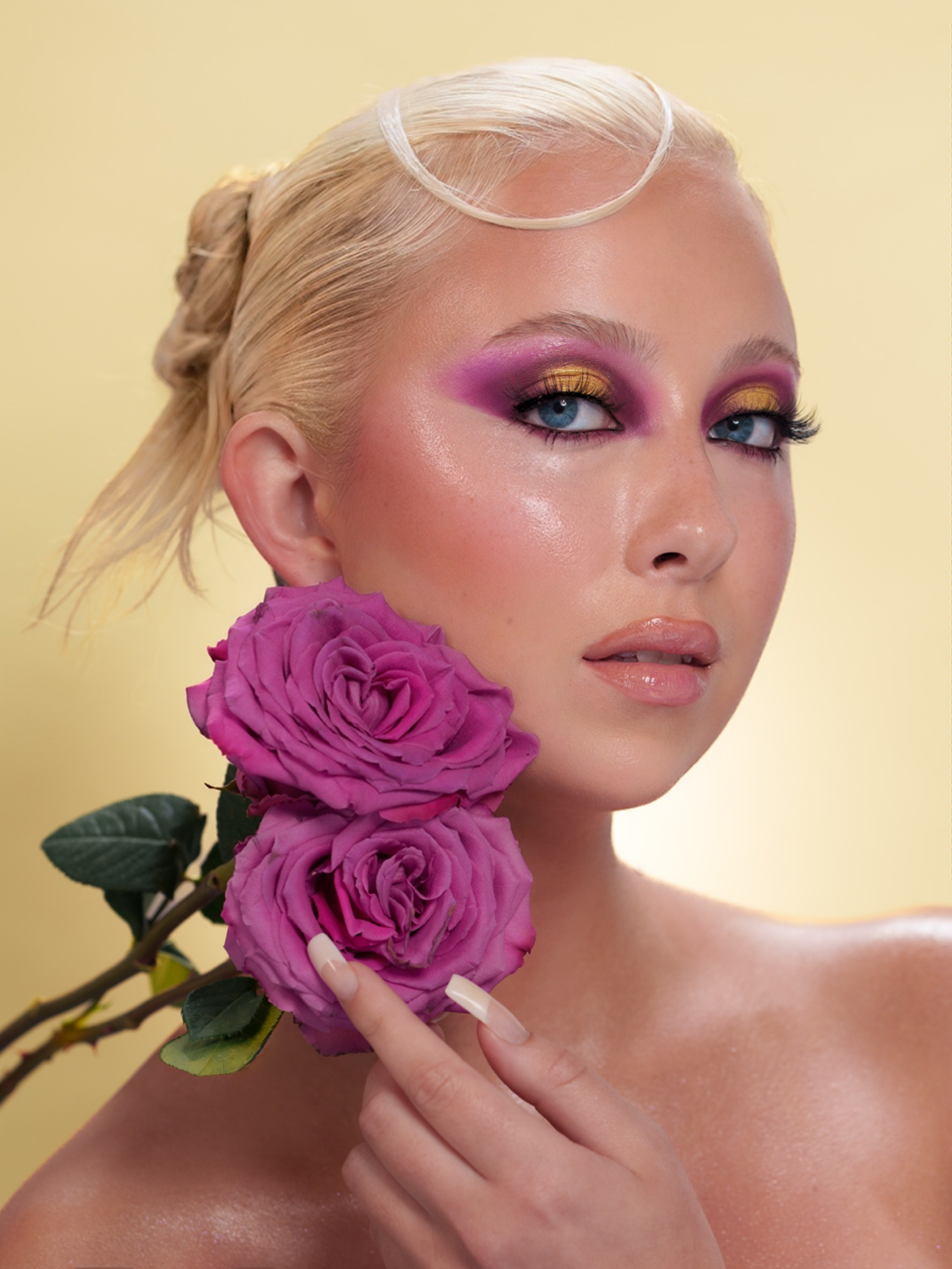 Blonde woman with bold purple and gold eyeshadow holds pink roses against a yellow background.