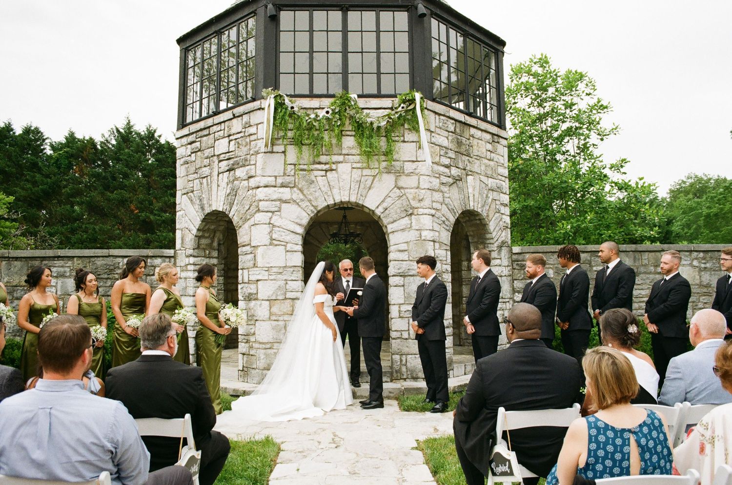 Outdoor wedding ceremony at a stone castle venue with bride, groom, and wedding party gathered under an arch.