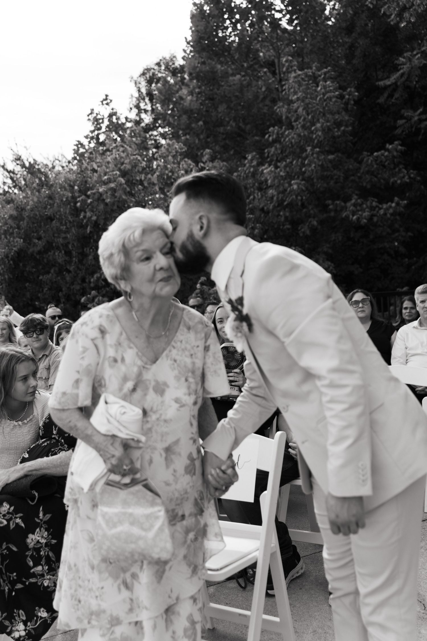 Groom in white suit kisses elderly grandmother on cheek at outdoor wedding ceremony with seated guests.