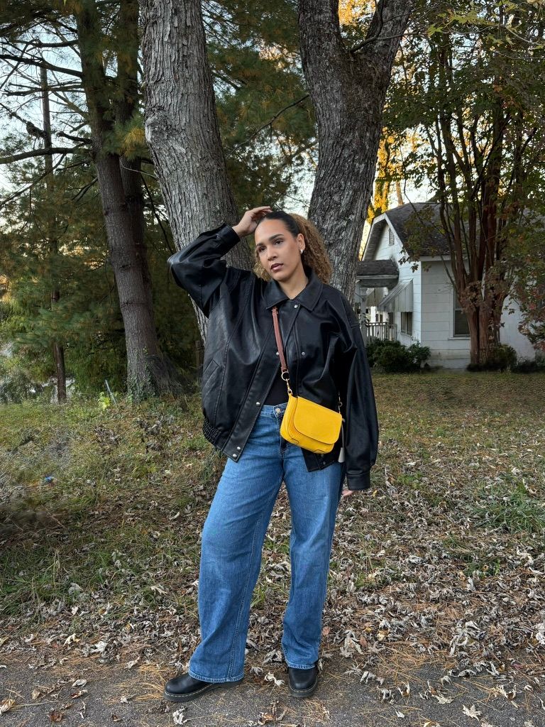 Woman in black leather jacket, wide-leg jeans, and yellow crossbody bag posing outdoors near trees.