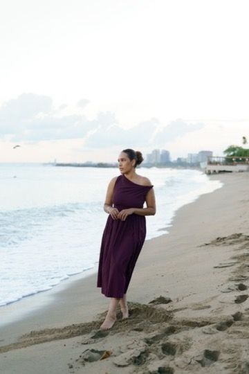 Woman in burgundy one-shoulder midi dress walking on sandy beach at dusk with city skyline in background.