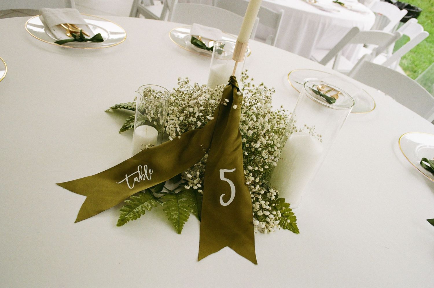 Wedding table centerpiece with baby's breath, greenery, and olive ribbon banner displaying table number 5.