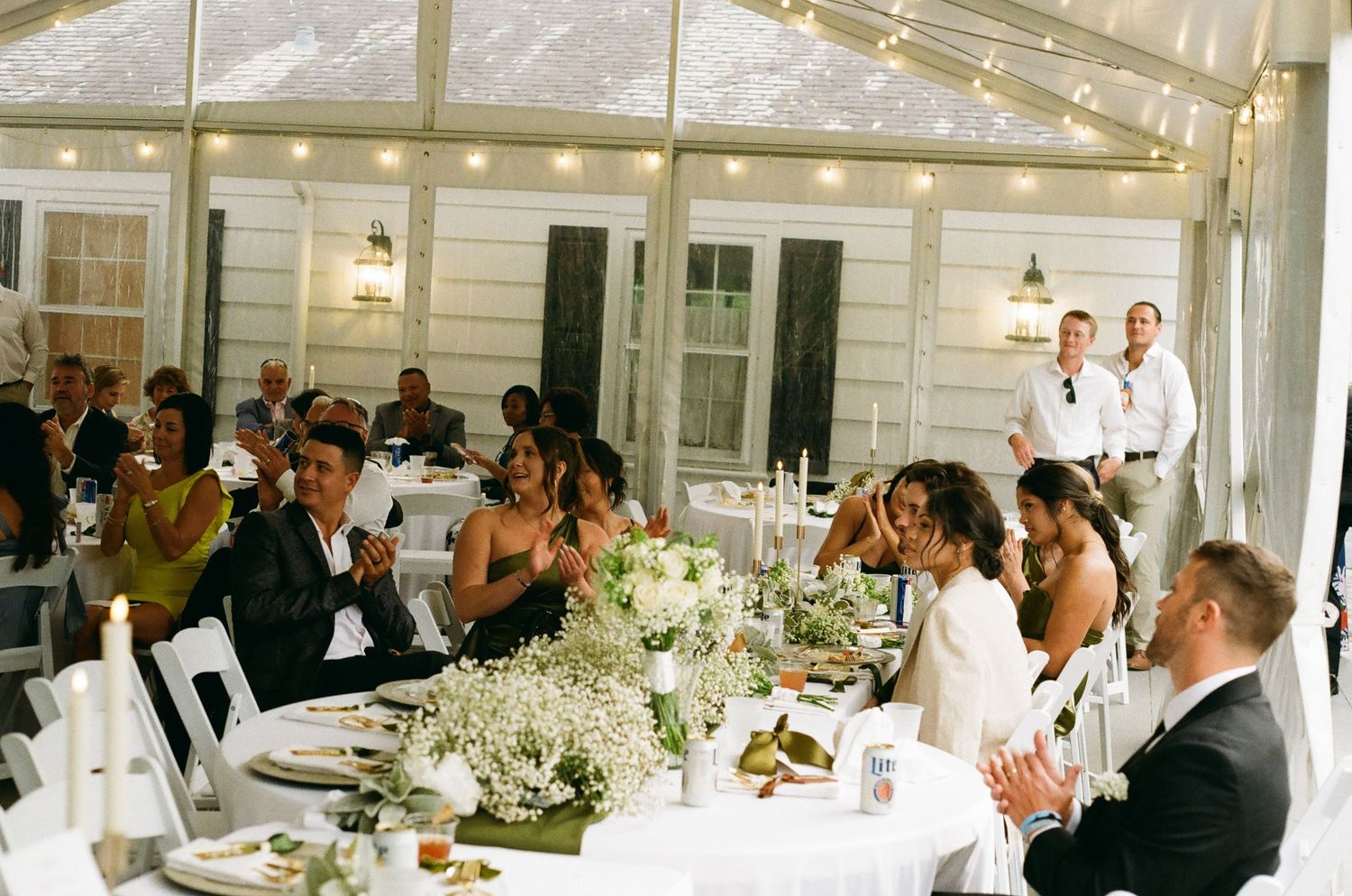 Elegant wedding reception dinner with guests seated at a long floral table under string lights in a white tent venue.