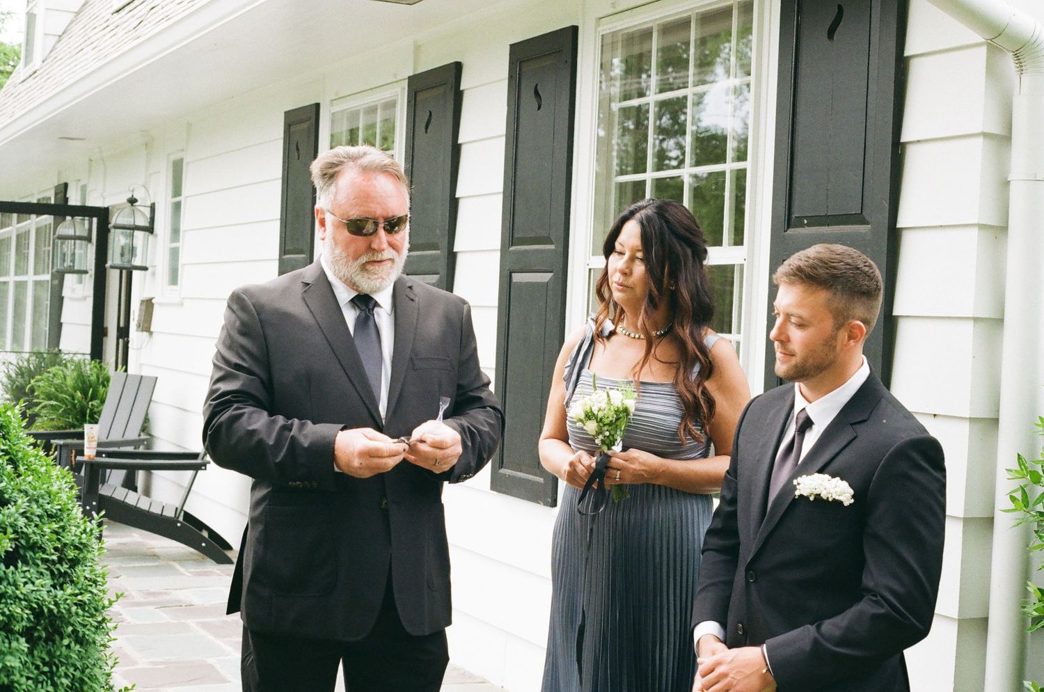 Three elegantly dressed people stand outside a white building, two men in suits and a woman in a gray gown holding flowers.