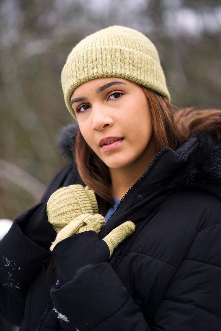 Young woman in green knit hat and matching mittens wearing black winter jacket outdoors in snowy setting.