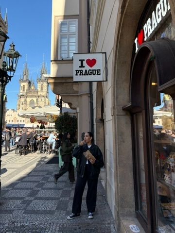 Woman standing beside an I Love Prague sign on a cobblestone street with the Church of Our Lady before Týn in background.