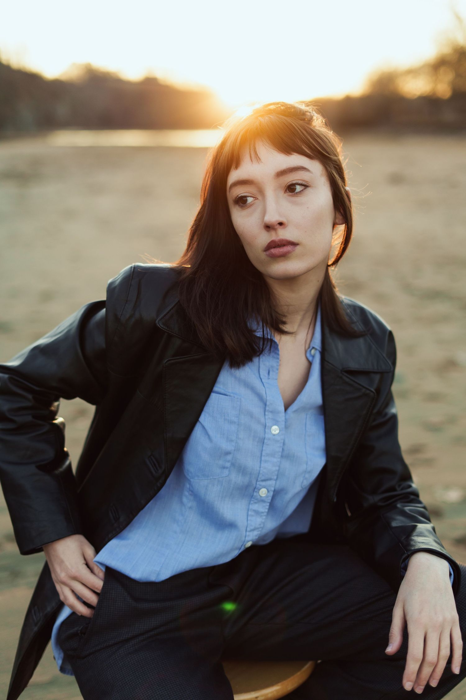 Model wearing a light blue button-down shirt and black leather jacket poses outdoors at sunset in a natural setting.