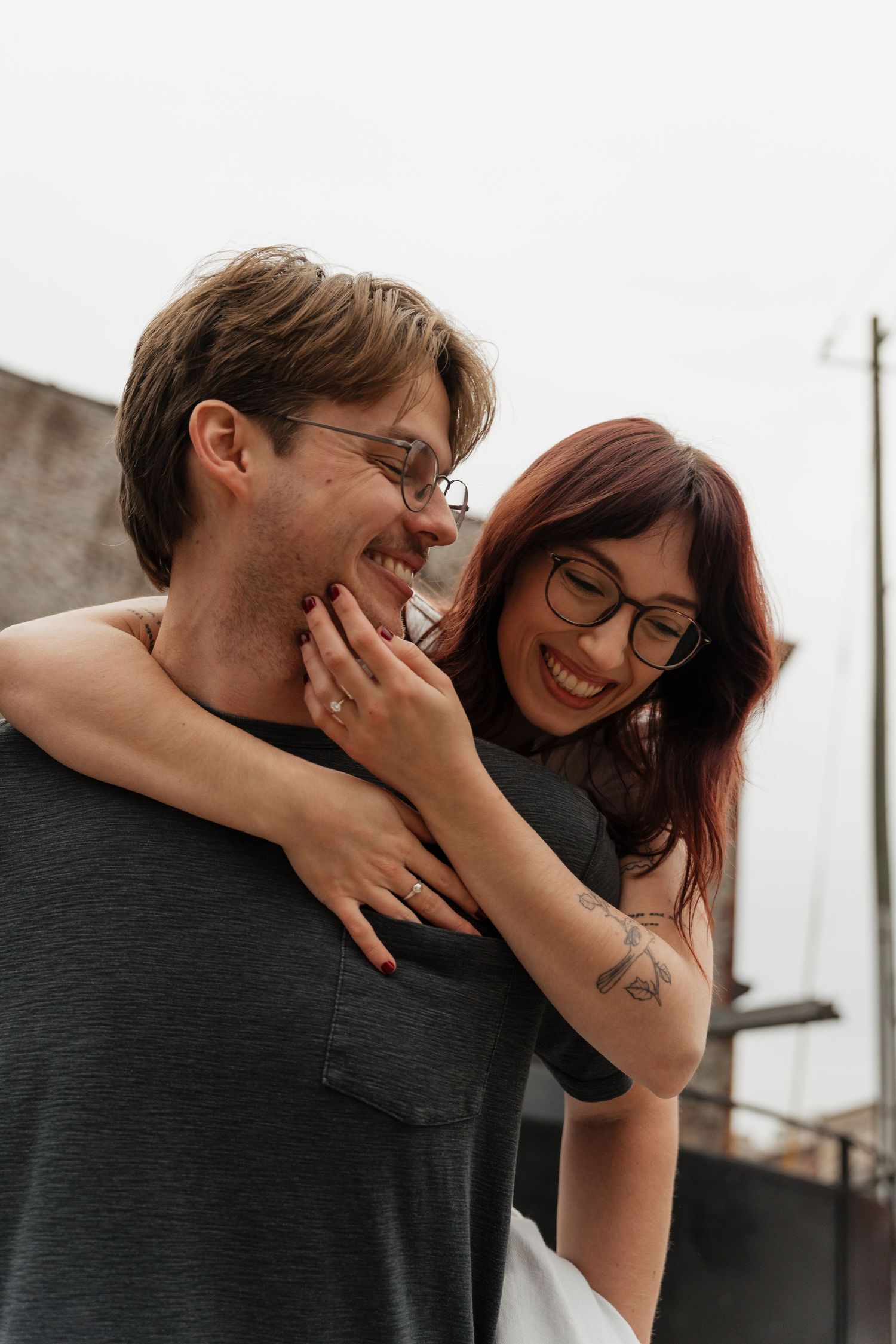 A smiling couple wearing glasses embrace outdoors, woman with red hair and tattoo hugs man from behind.