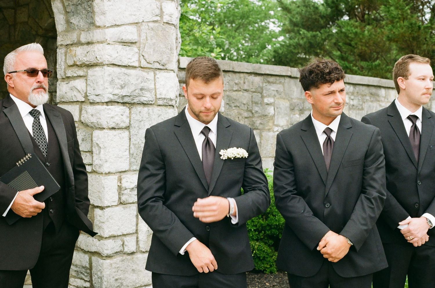 Groom and groomsmen in black suits standing outdoors during wedding ceremony, with stone pillar background.