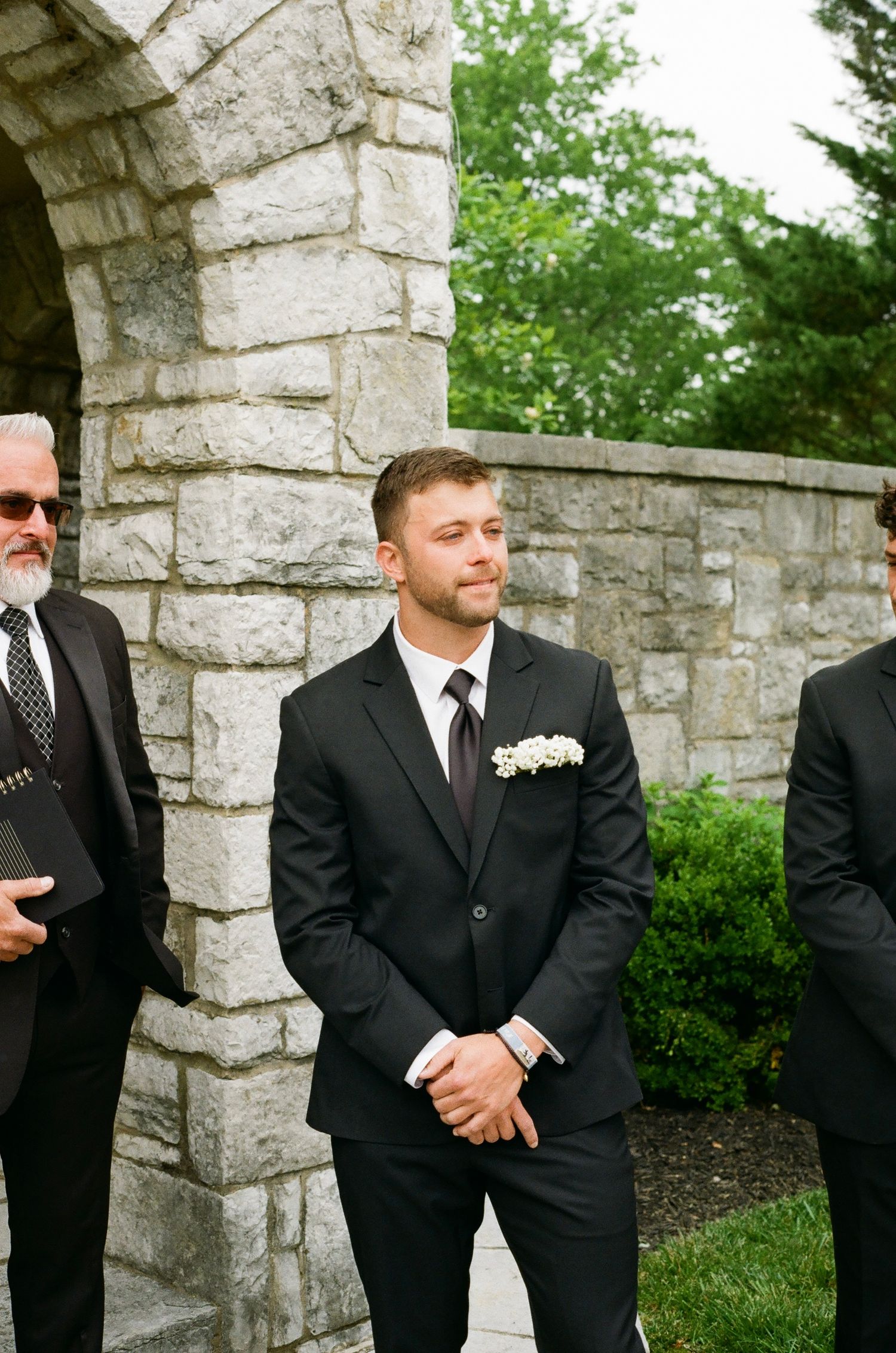 Groom in black suit with white boutonniere stands at outdoor stone wedding venue, emotional as bride approaches.