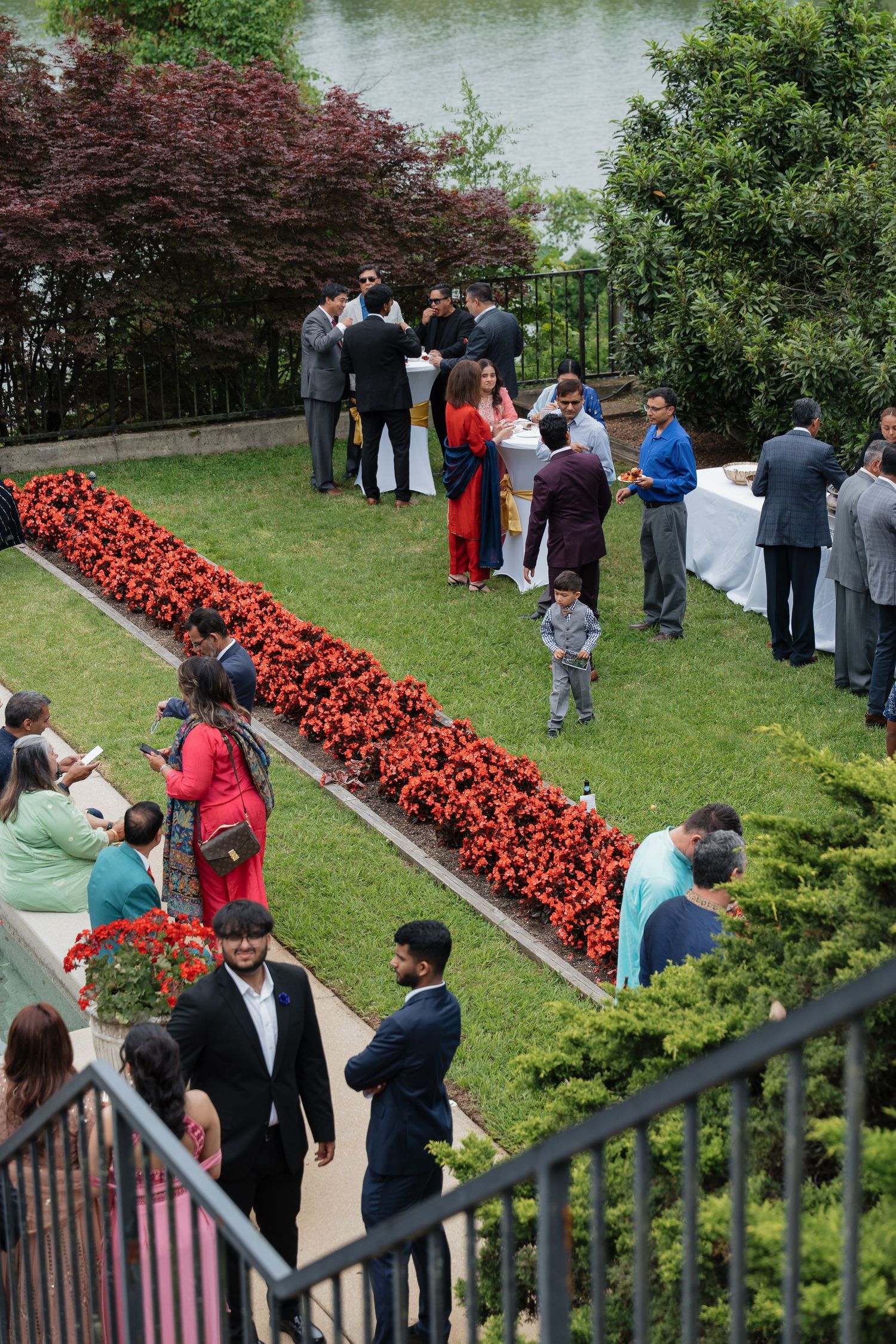 Outdoor garden party with guests mingling near red flower beds and a serene waterfront backdrop.