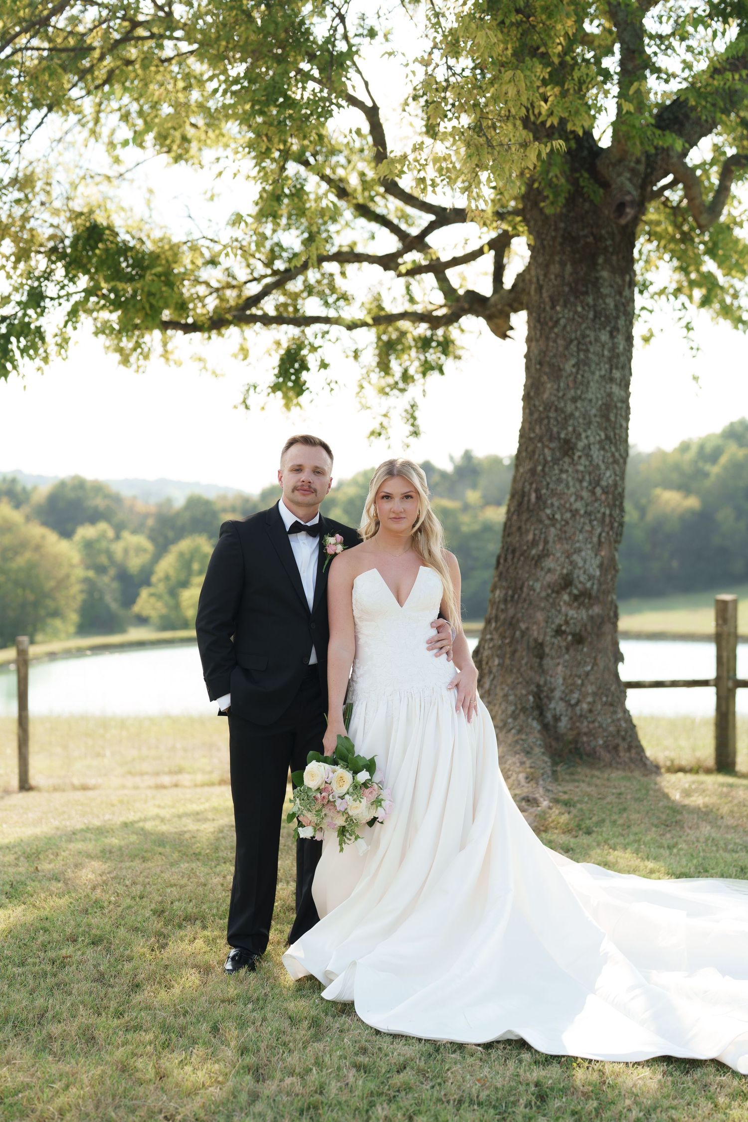 Bride in white gown and groom in tuxedo pose under large oak tree with pastoral pond views at outdoor wedding.