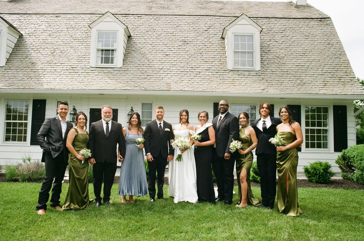 Wedding party poses in front of a white shingled house, bride in white gown with groomsmen and bridesmaids in olive green.