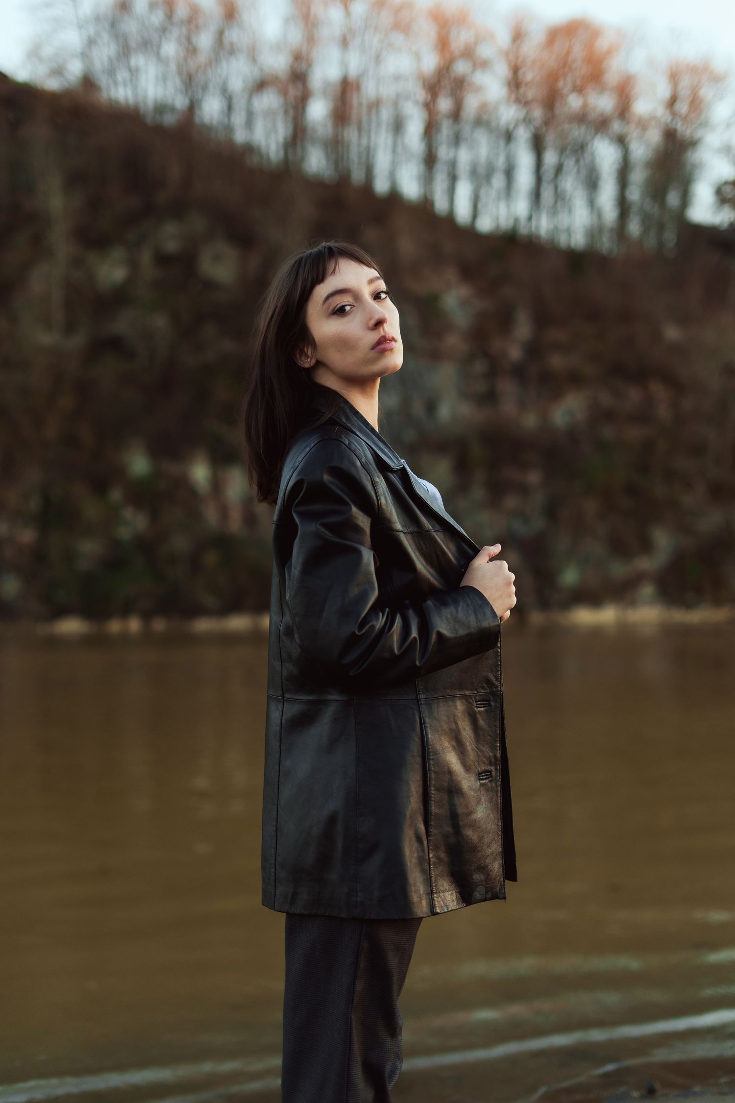 Artistic portrait of a person in a black leather coat posing thoughtfully by a calm lake during autumn.