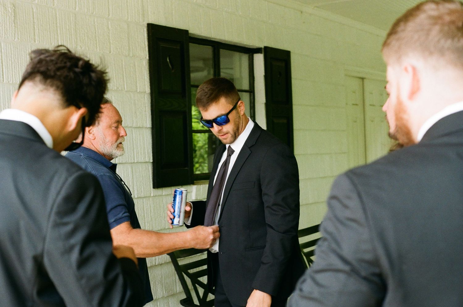 Men in black suits gathered on a porch, one wearing blue sunglasses, in a formal outdoor setting.