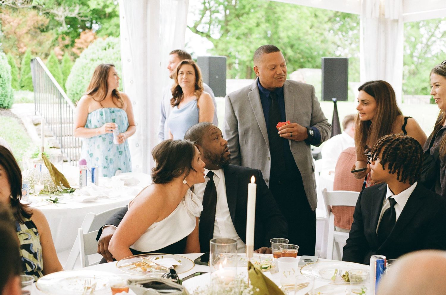 Guests mingle and converse at an elegant outdoor wedding reception under a white tent with floral centerpieces.