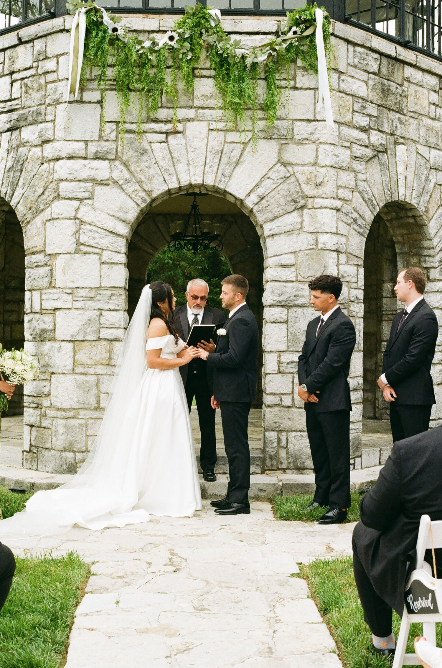 Outdoor wedding ceremony at stone chapel with bride in white gown, groom in black suit, officiant, and groomsmen on green lawn.