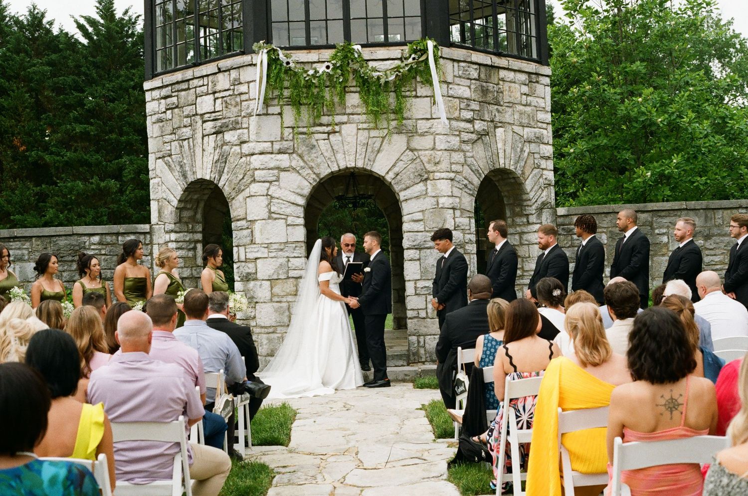 Outdoor wedding ceremony at a stone tower venue with bride and groom at altar, wedding party, and seated guests on a sunny day.
