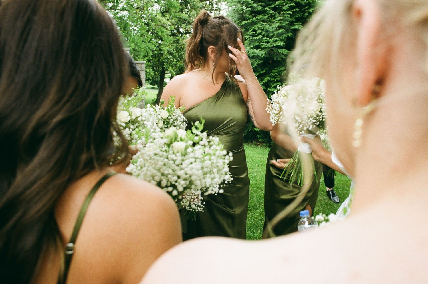 Bridesmaids in olive green dresses holding white floral bouquets gather outdoors in a lush garden setting.