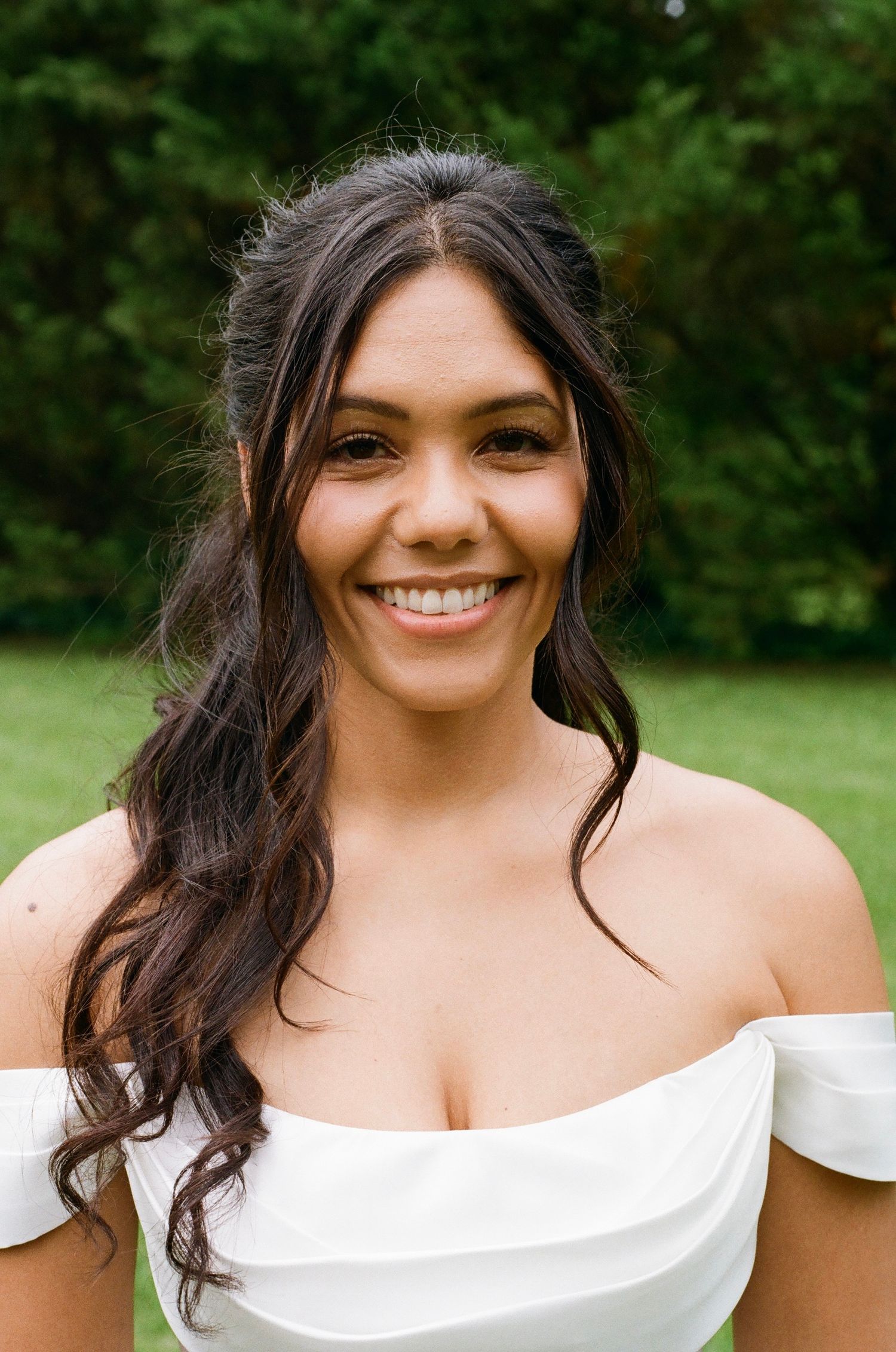 Smiling young woman with dark wavy hair wearing an off-shoulder white dress, posing outdoors with green trees behind her.