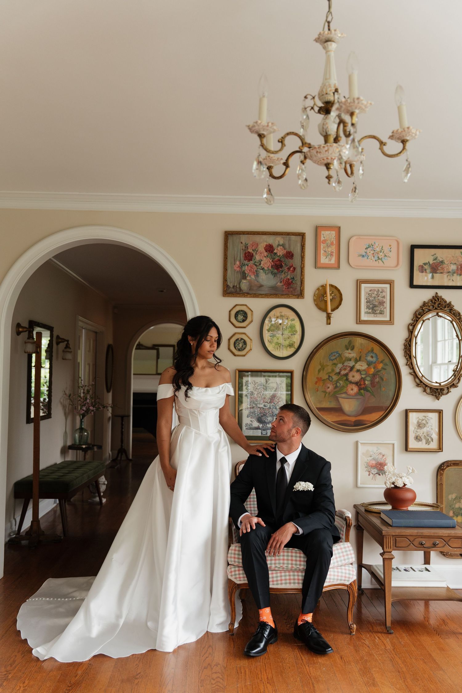 Bride in white off-shoulder gown stands beside groom in black suit seated on vintage sofa in elegant room with gallery wall.