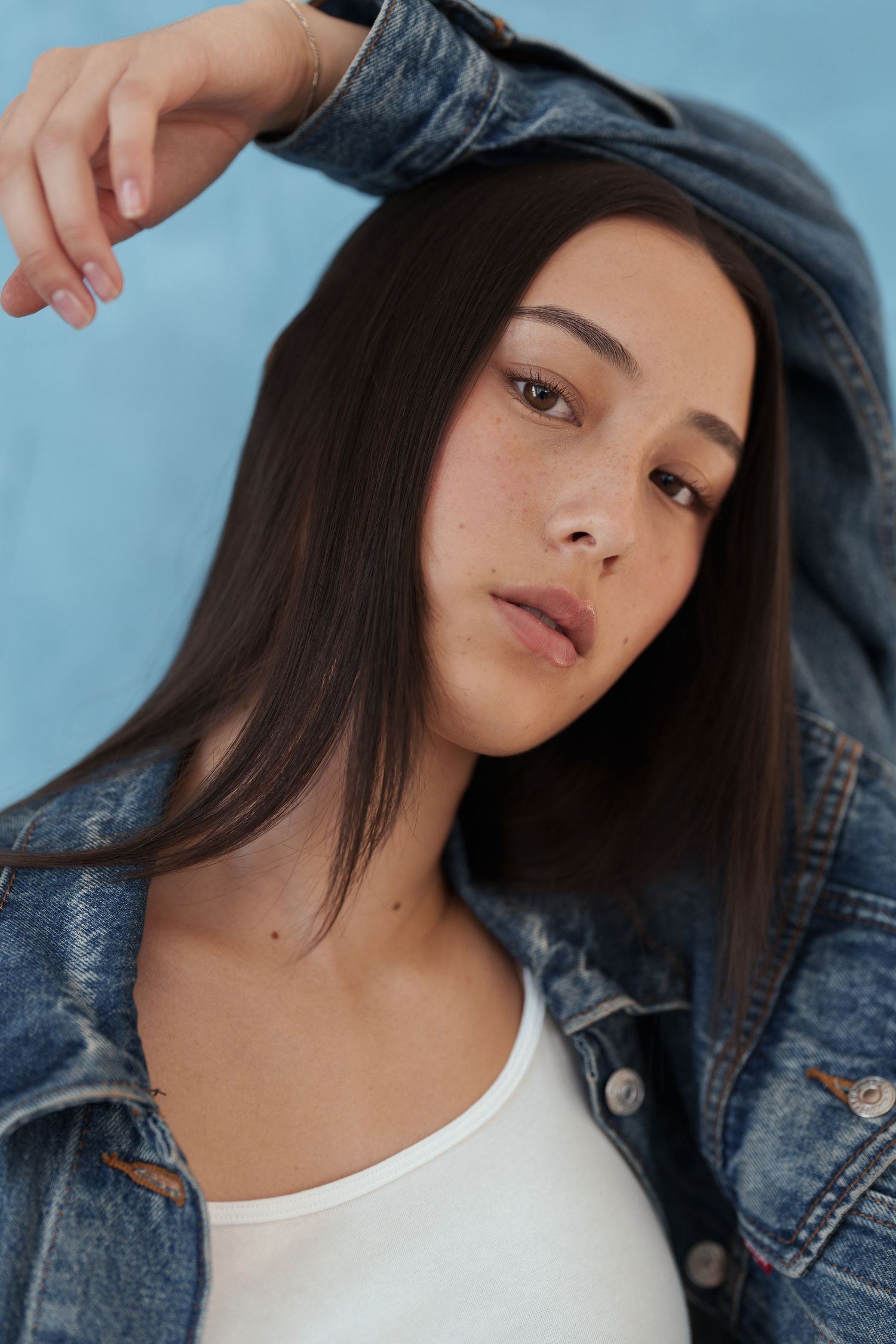 Young woman with long dark hair wearing a white tank top and denim jacket poses against a blue background.