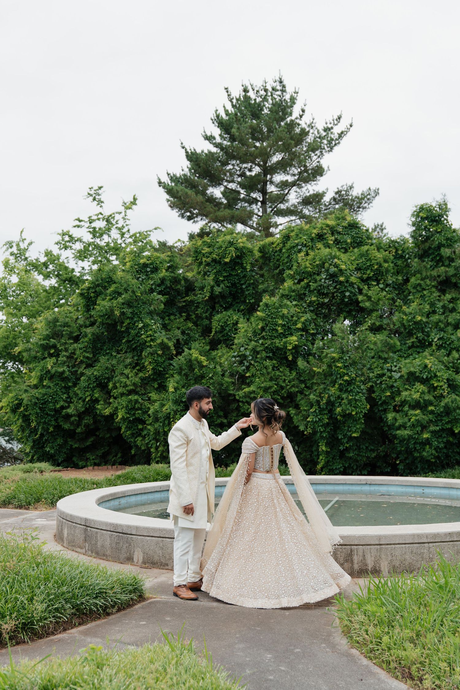 A couple in elegant cream-colored formal attire strolls along a curved concrete path surrounded by lush greenery.