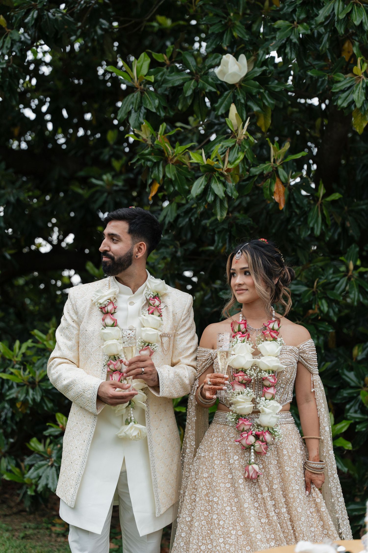 Elegantly dressed couple holding drinks outdoors with magnolia trees in background at wedding reception.