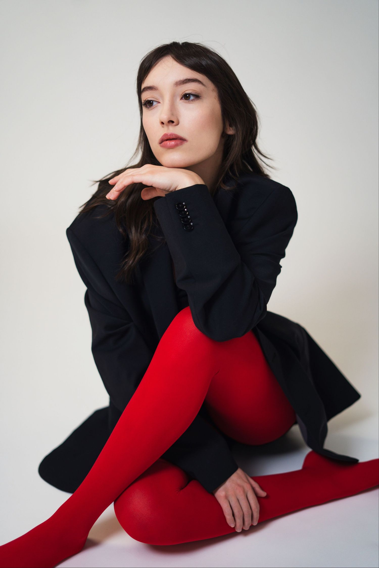 Woman in black blazer and red bodysuit poses dynamically against white backdrop in fashion photoshoot series.