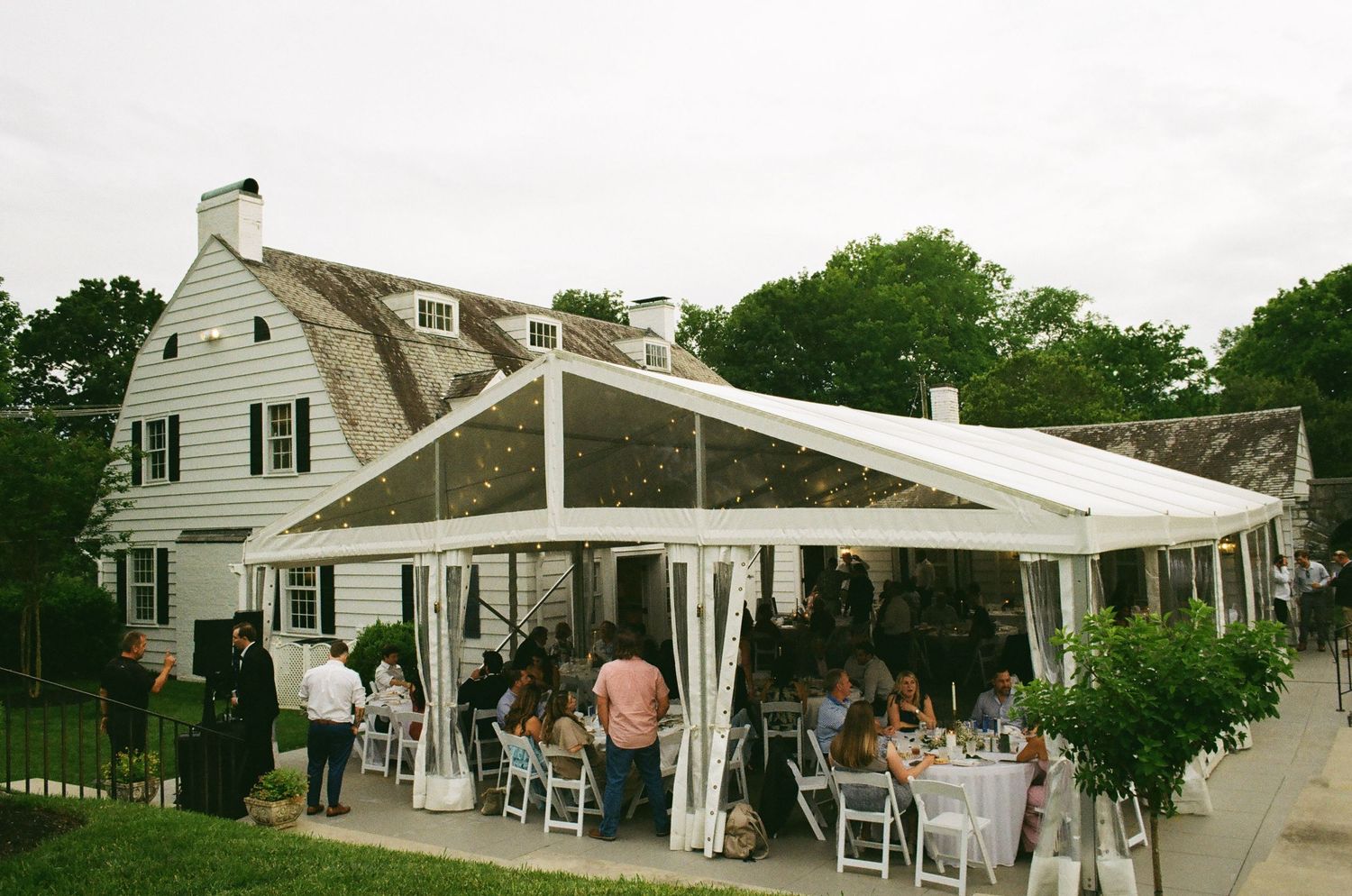 Elegant outdoor wedding reception under a white tent canopy beside a classic white colonial venue surrounded by lush trees.