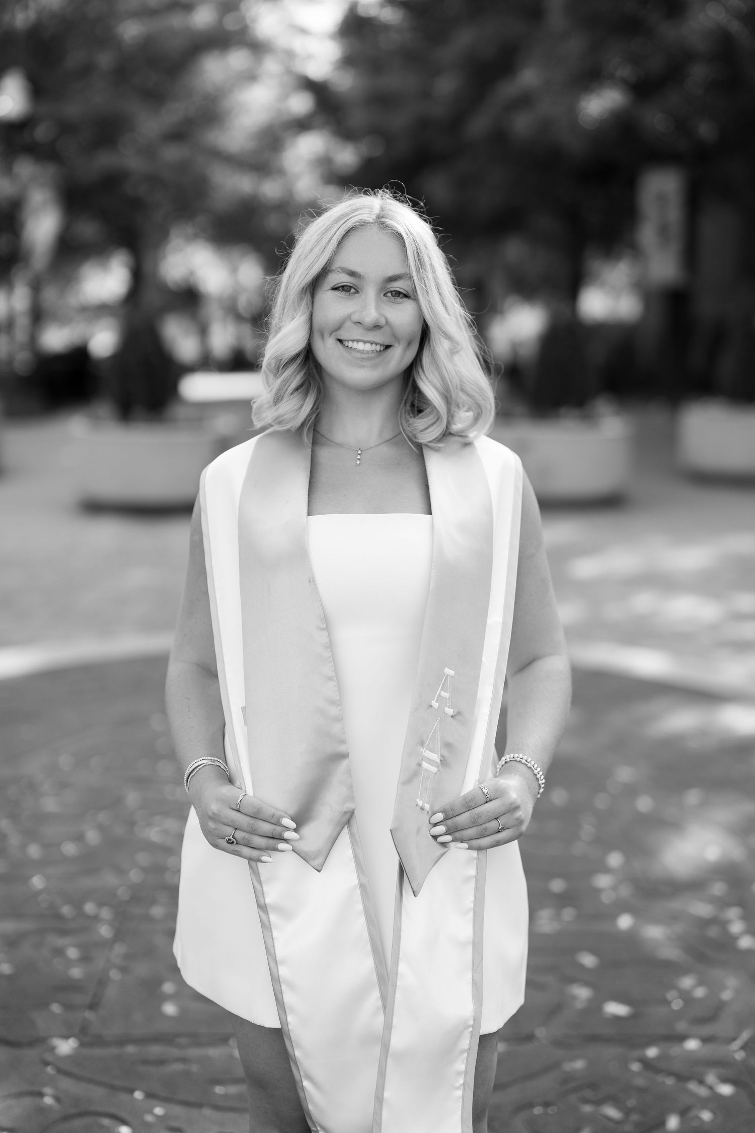 Smiling blonde woman in white dress wearing graduation stole poses outdoors on brick pathway.